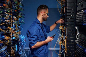Blue colored lighting. Young man is working with internet equipment and wires in server room