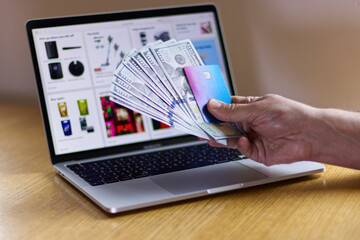 Man holding hundred dollar bills and credit card near laptop keyboard with online store displayed on blurred computer screen.