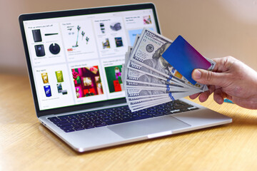 Man holding hundred dollar bills and credit card near laptop keyboard with online store displayed...
