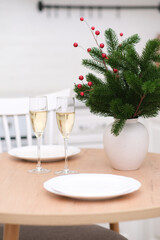 Two glasses of Champagne, two white plates on round wooden table. Christmas home interior. Ceramic vase with fir branches.