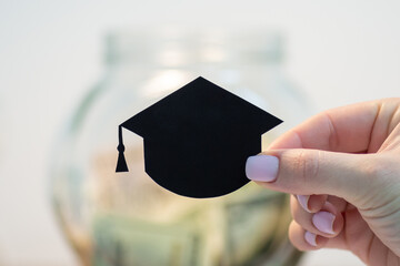 Close up of picture of small black students hat in female hand, money box on the background. Education, saving money and investment into the future concepts