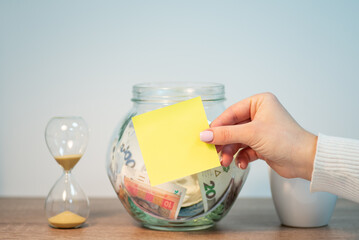 Female hand holding yellow paper sticker with copy space in front of glass money box. Ukrainian...