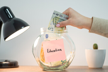 Five euro banknote in hand of a woman, who puts it into the transparent money box. Pink sticker with word Education on the glass jar, concept of saving money for future education