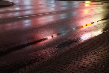 Nasse Straße mit bunter Lichterspiegelung zwischen Gehwegen in Stadt bei Wind, Regen und Kälte in...