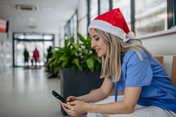 Beautiful nurse with christmas hat in hospital waiting area, holding smartphone, reading text...