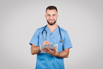 Smiling nurse writing on a clipboard in blue scrubs - obrazy, fototapety, plakaty