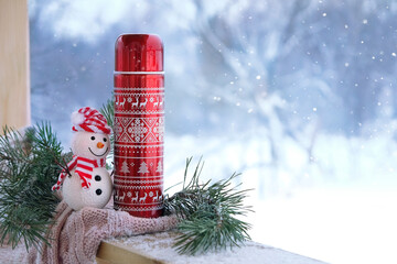 winter background. Snowman toy, Steel thermos with tea or coffee, knitted scarf, pine branch on...