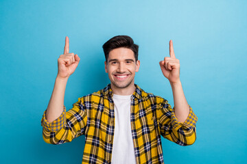 Photo of cheerful positive guy wear yellow checkered shirt pointing two fingers up empty space...