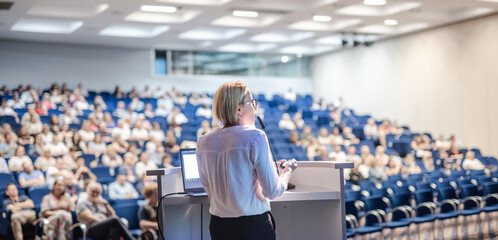 Female speaker giving a talk on corporate business conference. Unrecognizable people in audience at conference hall. Business and Entrepreneurship event - obrazy, fototapety, plakaty