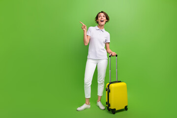 Full length photo of sweet excited girl dressed grey polo holding luggage pointing finger looking empty space isolated green color background