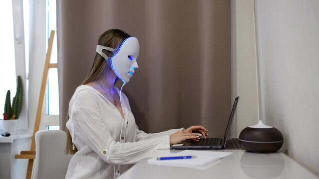 A beautiful girl with an LED mask on her head works at a laptop. Home skin care concept.