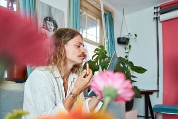 Young longhaired man with moustache holding lipstick and painting lips