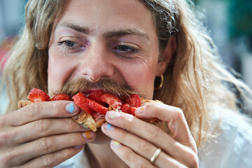 Creative cute man with makeup eating croissant with strawberry for breakfast