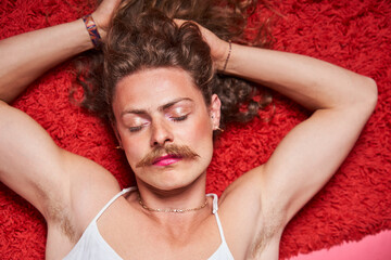 Longhaired man with makeup laying with closed eyes while posing - obrazy, fototapety, plakaty