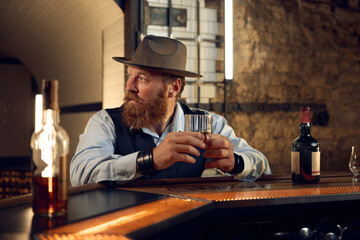 Portrait of mature gentleman in hat drinking alcoholic beverage in bar