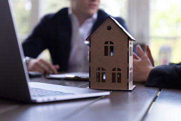 Potential client talks with estate agent about sale contract signing in agency office. Focus on wooden toy model of residence