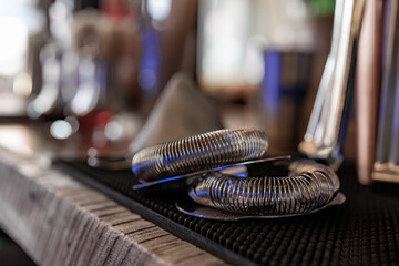 Metal, chrome, bar cocktail strainer sitting on a bar with a blurry background.
