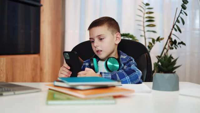 Concentrated school boy dressed in checked shirt sitting at table and watching videos on smartphone in bright room. Little boy having free time during distance learning with modern gadget in hands.