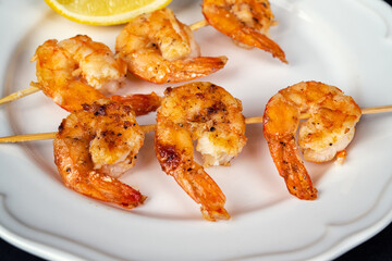 Delicious roasted shrimps on skewers arranged in a white plate with lemon slice and fresh rosemary on a black background close up