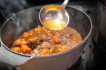 Clear broth is poured with a ladle into a casserole pot with steaming vegetables like carrot, onion, celery, leek and herbs, cooking a delicious sauce, soup or stew, copy space