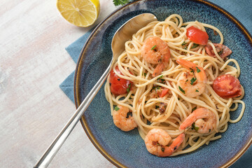 Spaghetti with shrimps, tomatoes and herbs in lemon sauce in a blue plate with spoon on a white wooden table, homemade Mediterranean seafood, copy space, top view from above