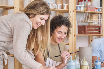Two joyful women share a laugh while painting on a ceramic piece in a bright, well-equipped pottery...