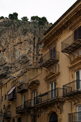 Old yellow residential house with few balconies in downtown of Cefalu. Typical Sicilian architecture. Rocky mountain with green trees on the top in the background. Travel and tourism concept. Sicily