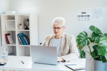 Concentrated grey hair middle-aged man working on laptop while sitting at his work place in home...