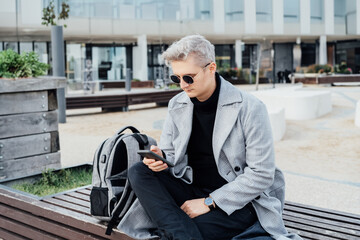 Young Handsome stylish man in elegant trendy coat and sunglasses checking message on mobile phone...