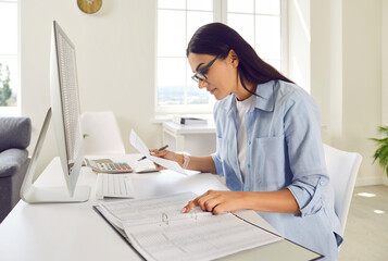 Portrait of a serious young business woman accountant in glasses sitting at the desk with pc computer with tables and charts in a light modern office on her workplace analyzing company data.