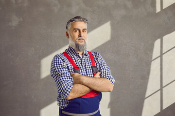 Senior repairman. Portrait of a mature male worker in uniform in the house. Bearded grey haired repair man in a shirt and blue workwear overalls standing with folded arms on a grey wall background