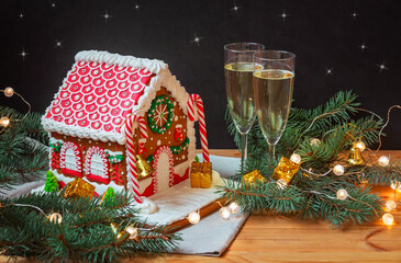 Gingerbread house with glaze standing on table with glasses of champagne, fir branches and glowing christmas lights.