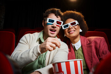 Obraz premium handsome man eating popcorn next to his jolly african american girlfriend on date at cinema