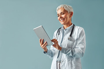 Beautiful mature female doctor in white coat holding digital tablet and looking at it, smiling while standing against blue background. Healthcare and digital technologies. - obrazy, fototapety, plakaty
