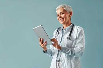 Poster Beautiful mature female doctor in white coat holding digital tablet and looking at it, smiling while standing against blue background. Healthcare and digital technologies. © HBS