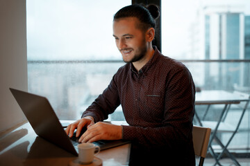 Portrait of young smiling man working on laptop at home, on background of panoramic windows.