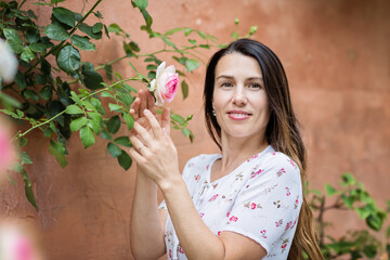 Lifestyle portrait of young stylish woman staying on the street in old town and touching rose flowers, wearing white dress