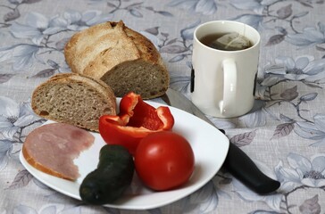 A plate containing cucumber, tomato, sweet pepper, a piece of ham and next to it a mug of tea with a focus on bread.