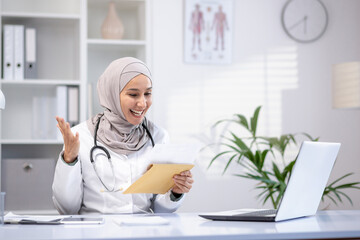 Happy female doctor in hijab holding letter in hands, sitting at table in hospital office, happy...