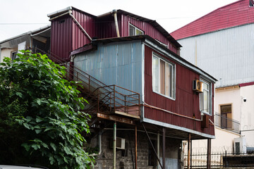 Charming elevated red and blue corrugated cottage on stilts, blending with lush foliage in Batumi's urban landscape, epitomizing summer in Georgia.