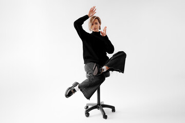 Fashionable young woman striking a dynamic pose on a chair, with a black outfit and bold boots, against a clean background