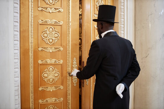 Back view of African American lord wearing top hat opening carved wooden doors and entering mansion, copy space