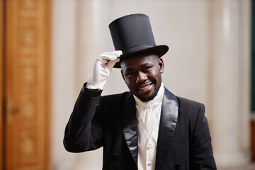 Waist up portrait of young Black gentleman wearing top hat smiling at camera in palace and giving...