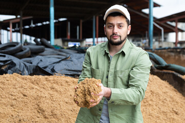 Interested young bearded farmer holding handful of brewers spent grains in open storage area at...