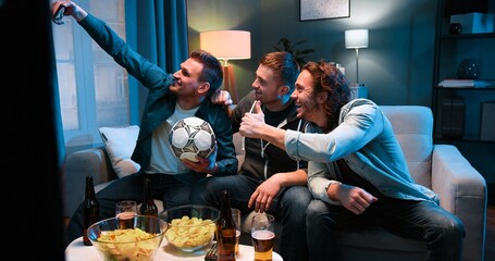 Fototapeta premium Three cheerful Caucasian friendly men sitting on sofa at night with football ball, snacks and beer and taking selfie photo with smartphone camera. Sport fans posing to phone in living room at TV.
