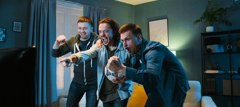 Happy Caucasian male football fans friends standing in front of TV screen in dark living room on vouch and watching match Euro2024. Scoring goal or winning game concept. Cheerful men celebrating win.