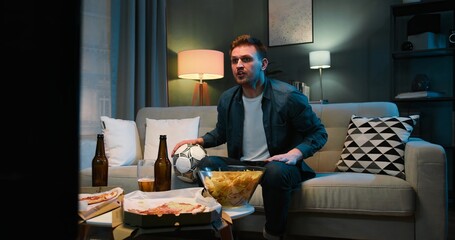 Caucasian young guy sitting on sofa at night in dark living room, eating potato chips and drinking beer. Male football fan is angry as favorite team loosing or missing goal. Man watching TV - obrazy, fototapety, plakaty