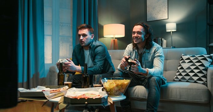 Handsome Caucasian joyful men friends sitting on sofa in tension and worrying while playing videogame with joysticks in front TV screen. Videogaming at home concept.