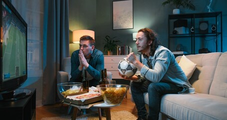Two male best friends cheering for football team in dark living room with snacks and beer. Caucasian men sitting on couch and worrying for game results. Guys watching sport channel on TV. - obrazy, fototapety, plakaty