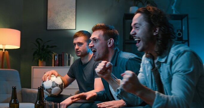 Tensed Caucasian male football fans screaming, worrying and cheering for favorite team while watching TV and sitting late at night in living room. Guys friends having nice evening together at home.